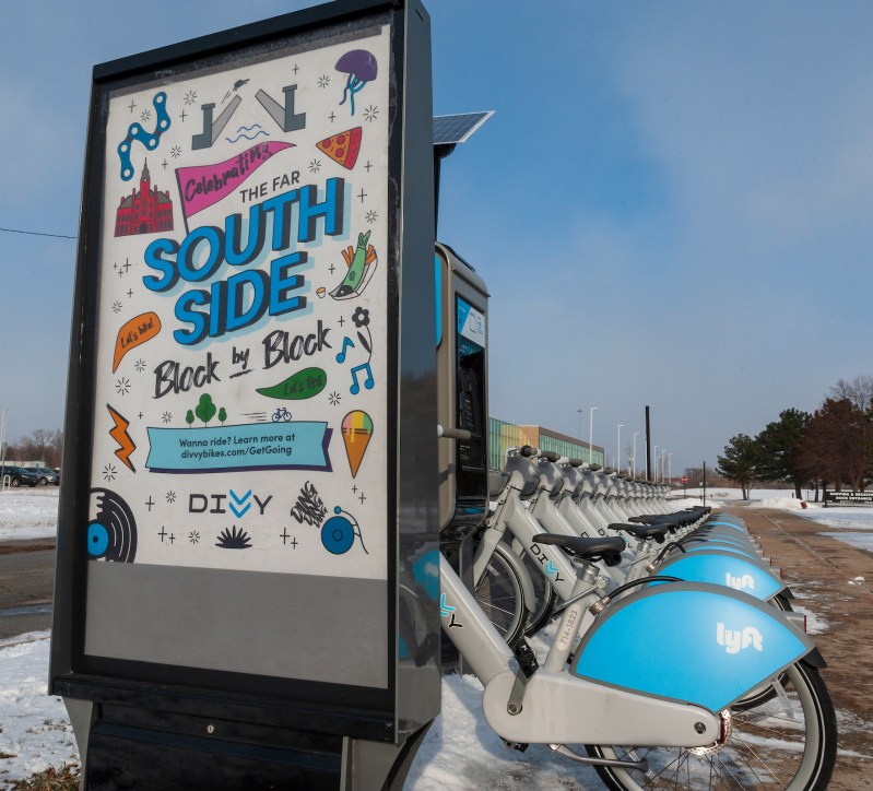 The new Divvy station at Olive-Harvey College, with the system's new model of electric bikes. Note the Blue Brothers car jumping the 95th Street bridge over the Calumet River at the top of the ad panel. Photo: CDOT